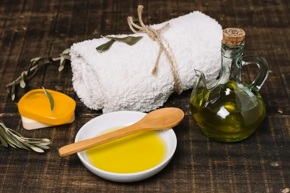 A shallow dish of golden oil with a wooden spoon across it, beside a glass cruet of olive oil, a rolled white towel, a bar of soap and olive sprigs on dark wood.