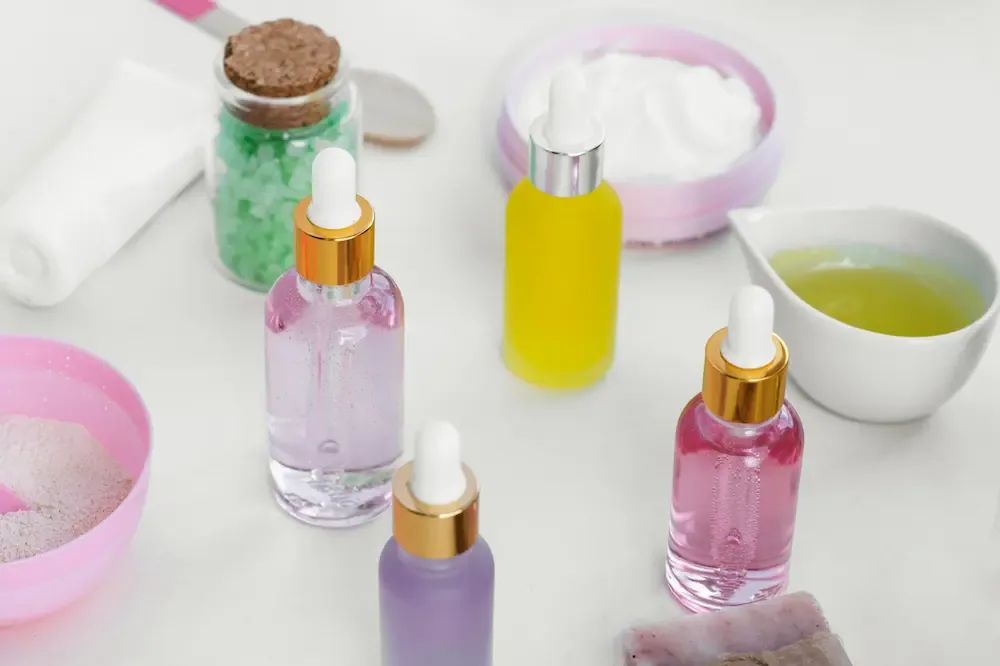 Four dropper bottles of serum in pink, purple and yellow on a white surface, with open pots of cream and powder and a small bowl of pale green oil.